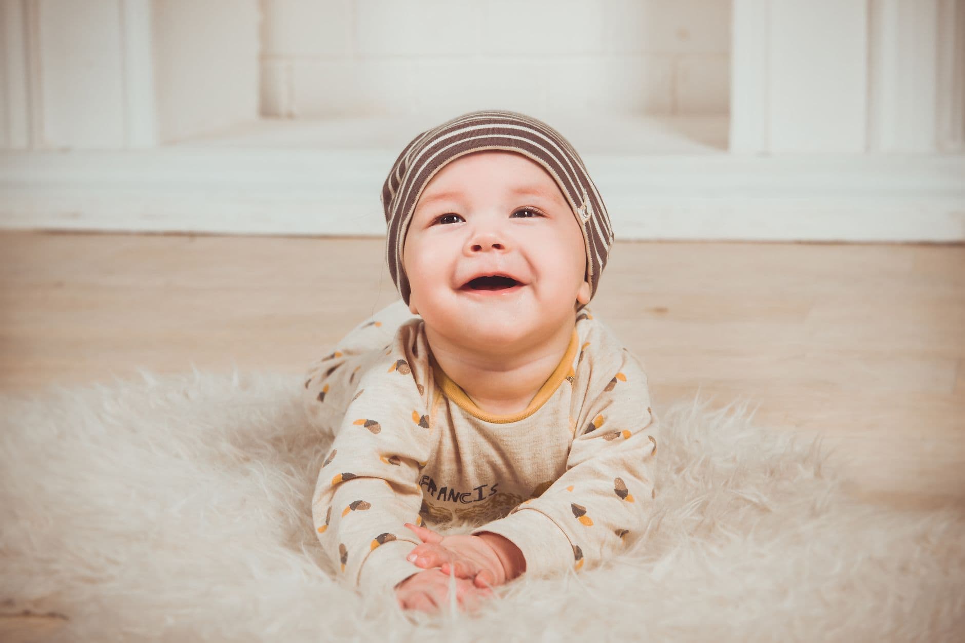 A baby in a cream patterned outfit and striped hat, laughing, propped up on a soft white rug